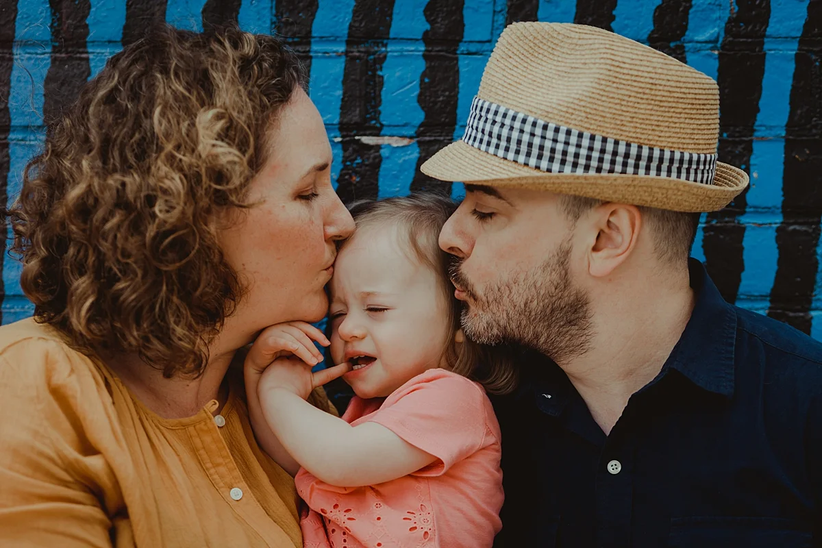 photo of dad and mom kissing youngest daughter in front of bright and colorful graffiti wall in dumbo brooklyn. photo by nyc family and newborn photographer krystil mcdowall