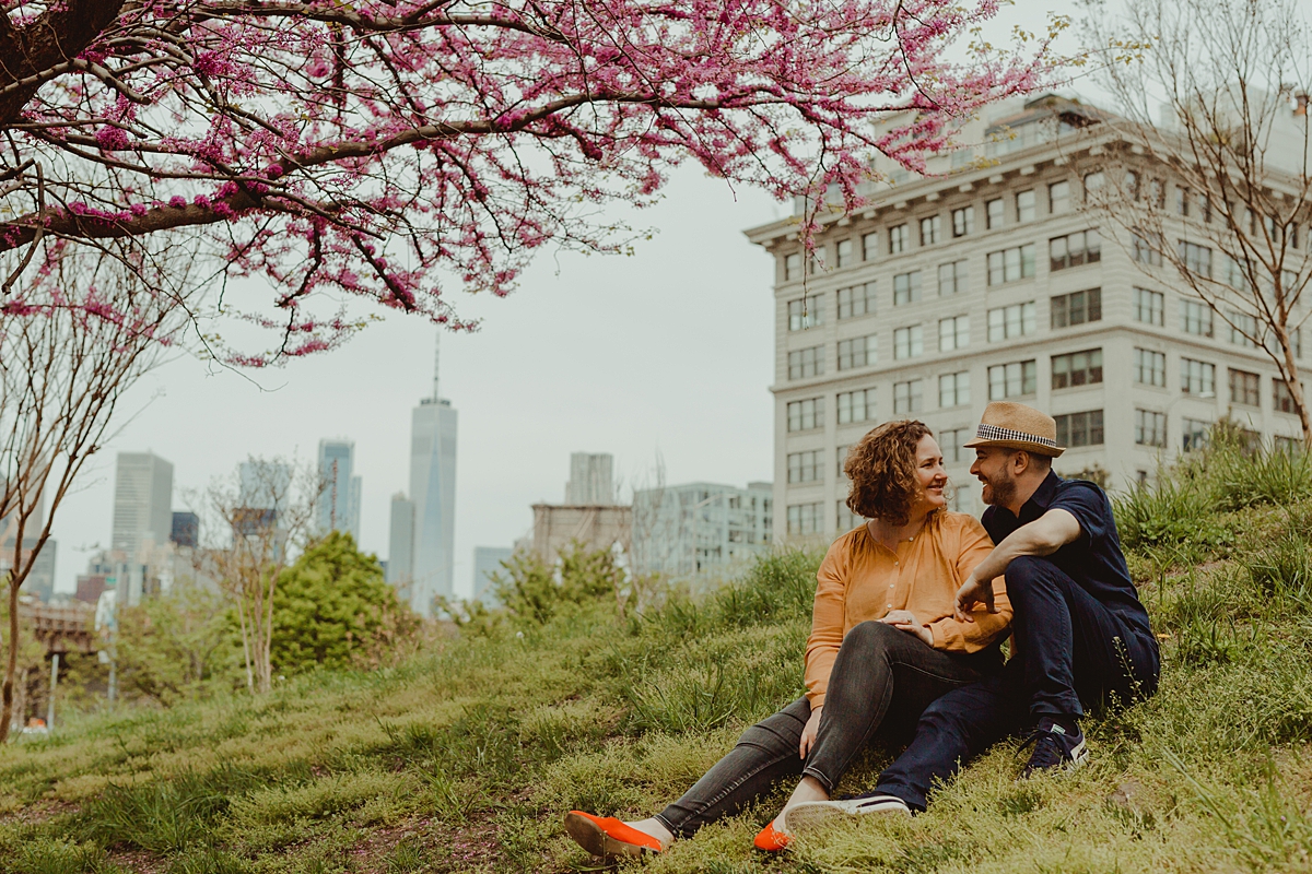 portrait of mom and dad sitting on green hill with cherry blossoms overhanging and manhattan buildings in the background. photo by nyc family and newborn photographer krystil mcdowall