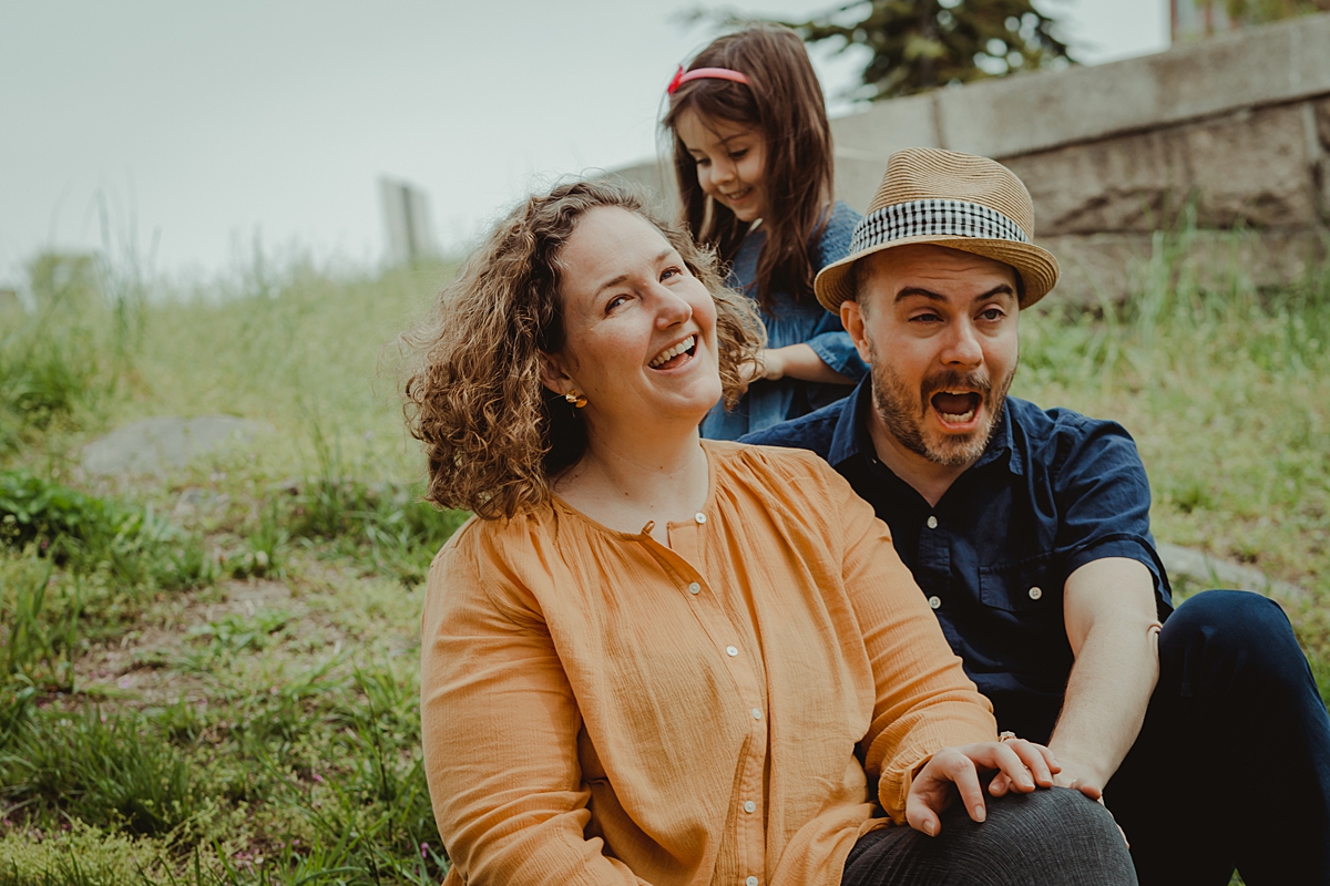 candid photo of young girl and mom and dad sitting on green grass amidst blooming cherry blossoms in brooklyn. photo by nyc family and newborn photographer krystil mcdowall