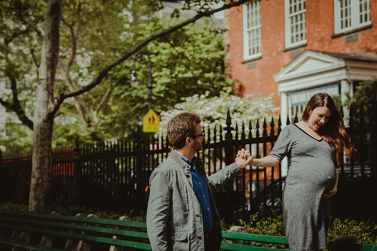 image of pregnant husband and wife in the park with pregnant mom walking along park bench while her husband holds her hand during nyc outdoor maternity photo session. photo by nyc family and newborn photographer krystil mcdowall