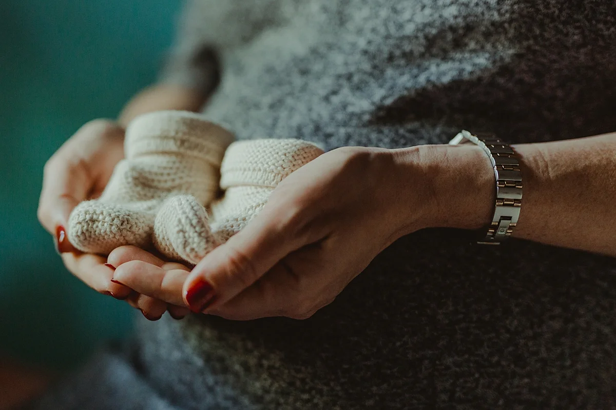 close up portrait of expecting mother holding white knitted baby shoes in front of her pregnant belly during maternity photo session. photo by nyc family and newborn photographer krystil mcdowall
