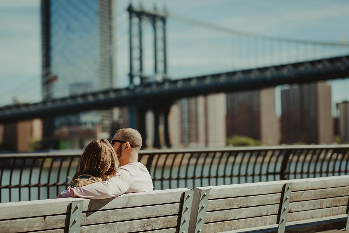 photo of pregnant husband and wife sitting on park bench in brooklyn bridge park looking at the manhattan bridge during outdoor maternity photo session in brooklyn. image by krystil mcdowall photography