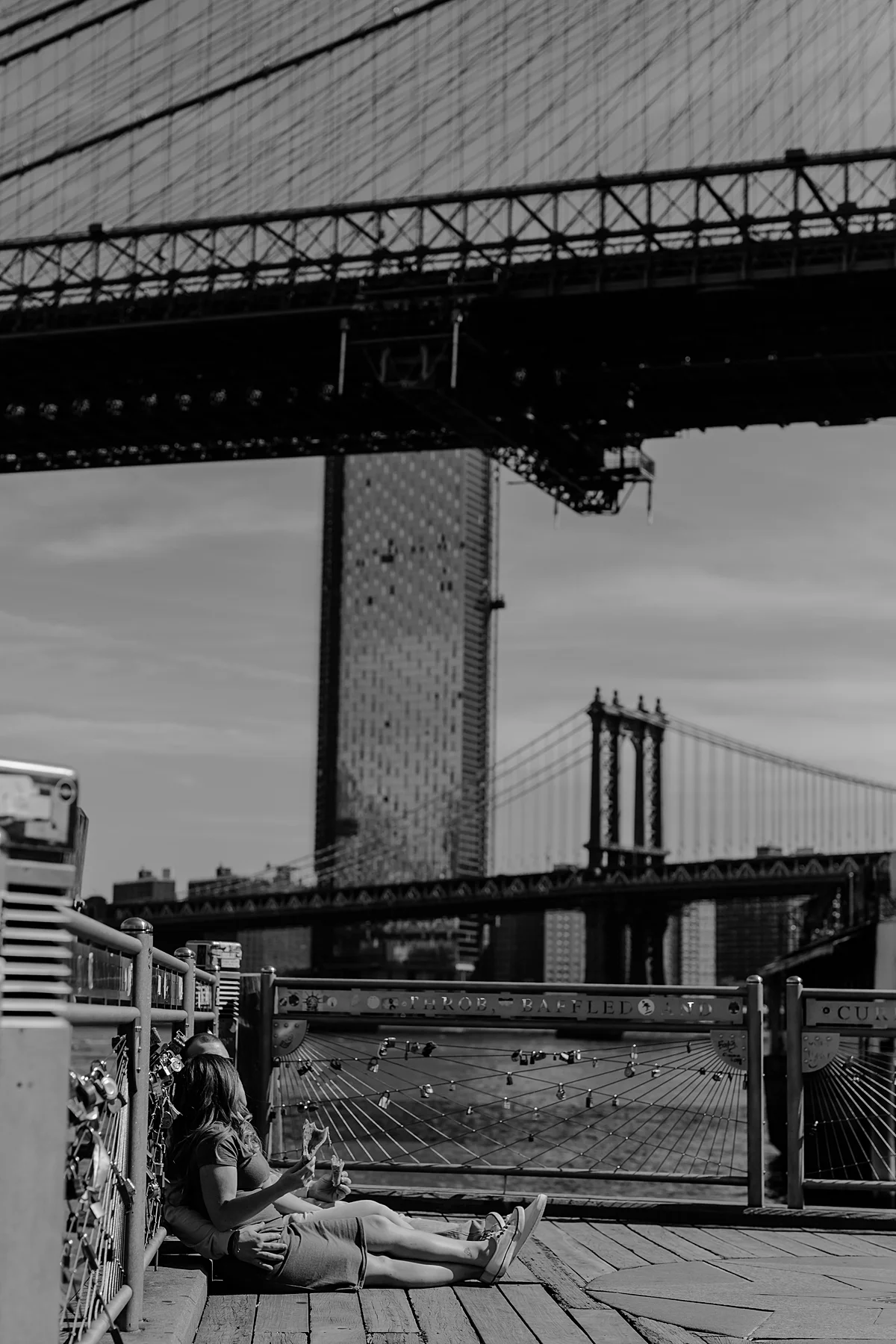 black and white photo of pregnant couple laughing and eating pizza while sitting on the pier at brooklyn bridge park during maternity photo shoot. image by krystil mcdowall photography