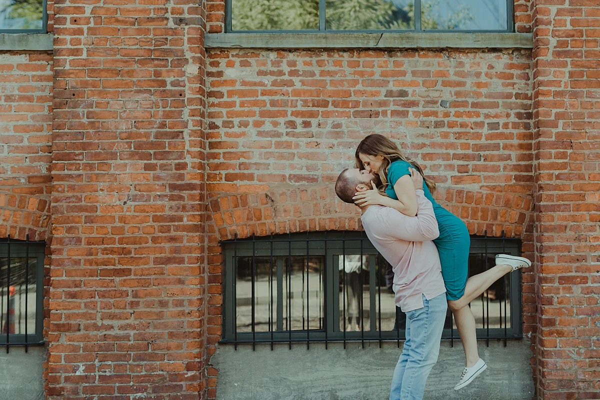 color image of husband and wife kissing in front of gritty brick wall in dumbo brooklyn during maternity photo shoot. image by krystil mcdowall photography