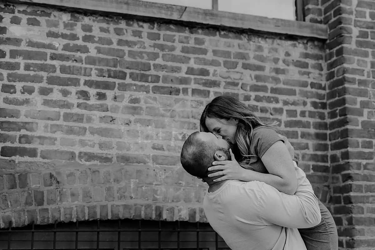 husband and wife laughing and kissing in front of gritty brick wall in dumbo brooklyn during maternity photo shoot. image by krystil mcdowall photography