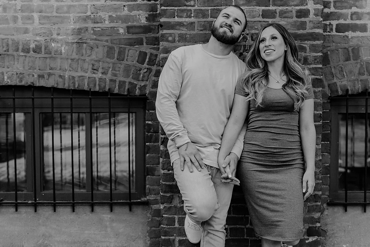 black and white image of husband and wife looking out at the manhattan skyline while leaning again gritty brick wall in dumbo brooklyn during maternity photo shoot. image by krystil mcdowall photography