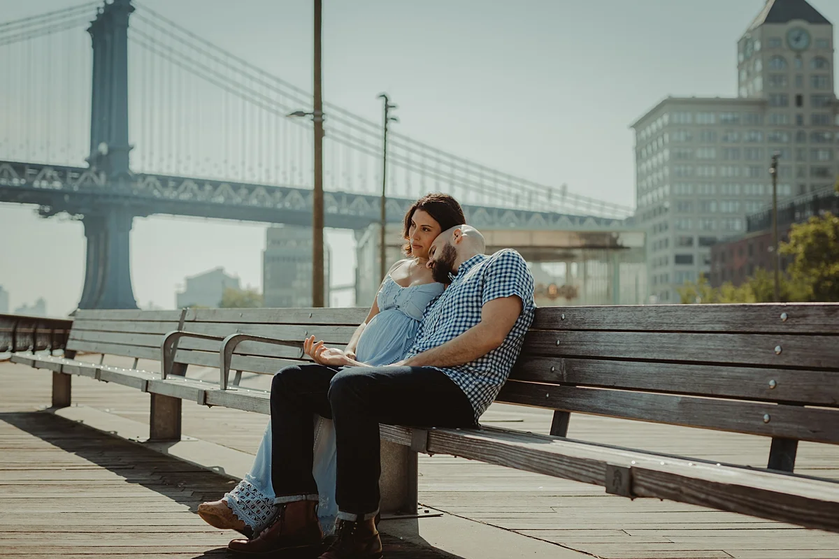 candid portrait of pregnant married couple sitting on park bench together looking out at the river and the city during lifestyle maternity photo session with orange county family photographer krystil mcdowall