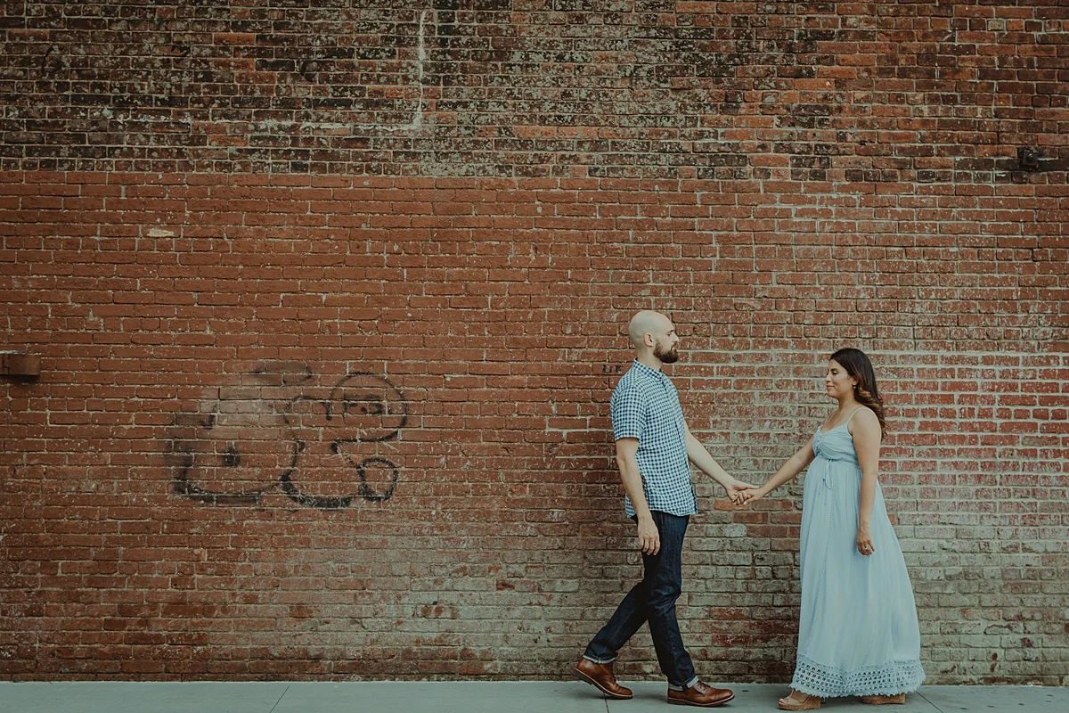 candid image of pregnant married couple walking along the street with gritty brick wall in the background during lifestyle maternity photoshoot. image by orange county family photographer krystil mcdowall