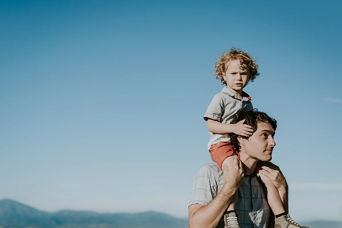 photo by Orange County family photographer Krystil McDowall. Photo of dad giving his cute blonde curly haired son a shoulder ride with mountains in the background during outdoor family lifestyle photo session at Top of the World Laguna Beach California
