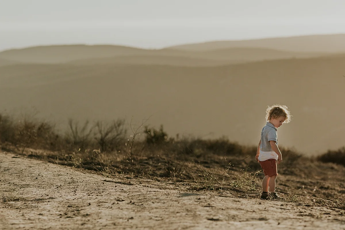 photo by Orange County family photographer Krystil McDowall. Candid photo of sweet boy in red shorts with curly blonde hair exploring the dirt and the mountains as the sun sets during family lifestyle photo session at Top of the World Laguna Beach California