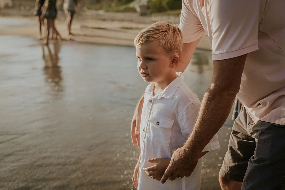 orange county family photographer. photo of dad holding his cute blonde son as ocean wave comes into the shore during outdoor family photo session at Corona del Mar State Beach with Krystil McDowall Photography
