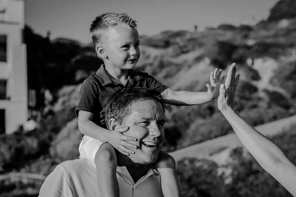 orange county family photographer. photo of dad giving his son a shoulder ride while he high 5's his mom during outdoor family photo session at Corona del Mar State Beach with Krystil McDowall Photography
