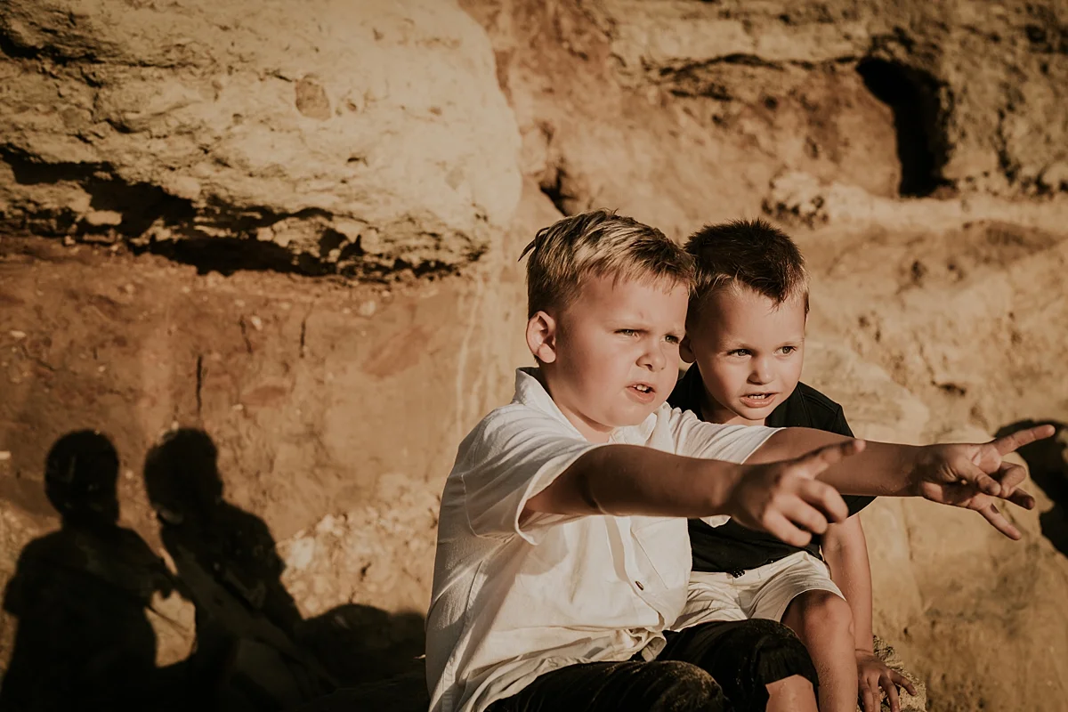 orange county family photographer. candid photo of two brothers sitting on rock bluffs at the beach and laughing and being silly during outdoor family photo session at Corona del Mar State Beach with Krystil McDowall Photography