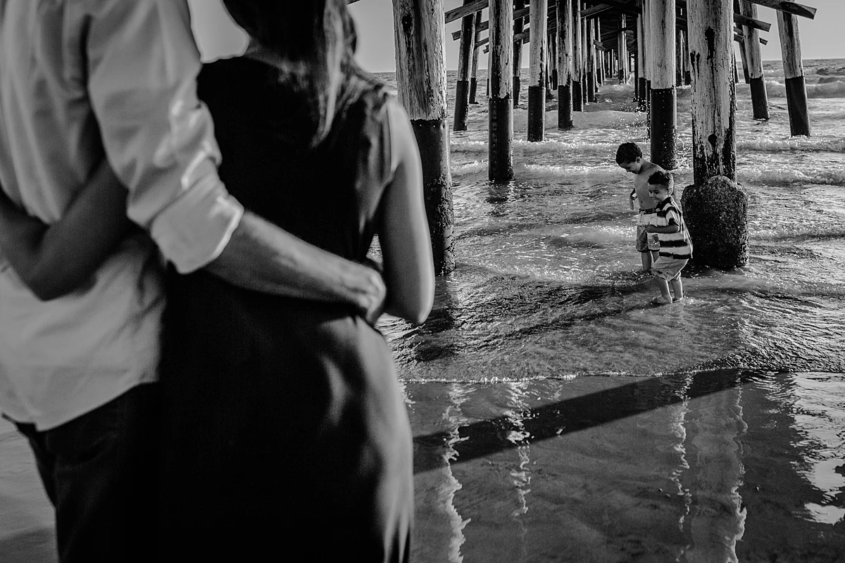 Orange County family photographer. Candid black and white photo of boys playing underneath the pier and in the ocean water while mom and dad look on at Huntington Beach California