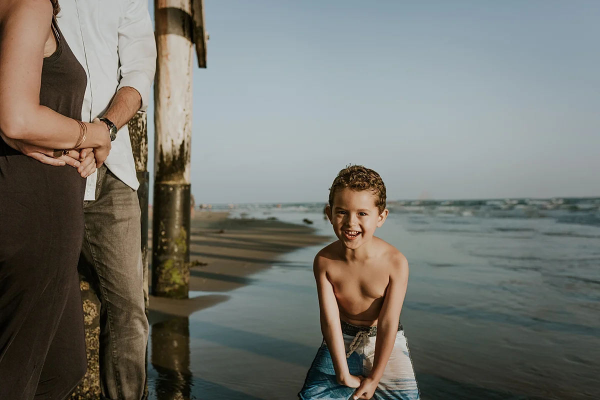 Orange County family photographer. Candid photo of son playing in the ocean water while mom and dad look on at Huntington Beach California