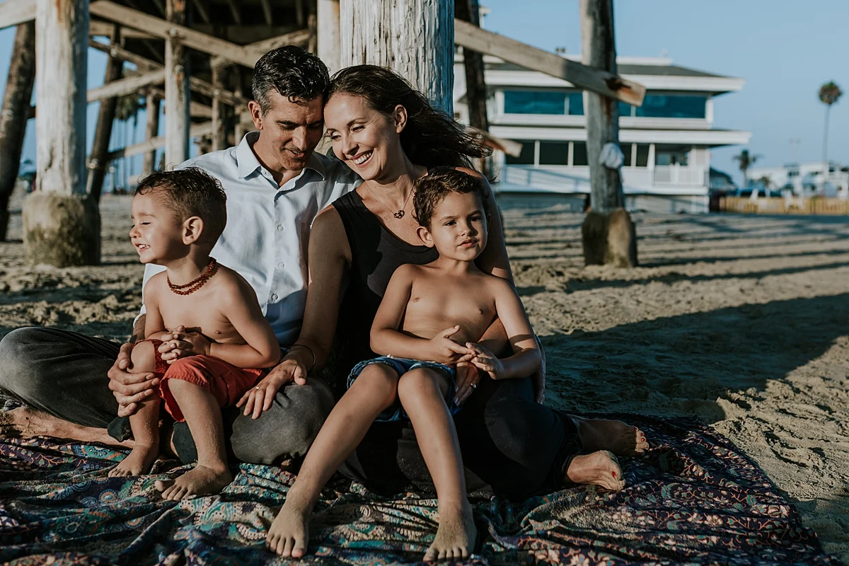 Orange County family photographer. photo of family of four sitting underneath the pier on beautiful detailed beach blanket during outdoor family photo session at Huntington Beach Pier California