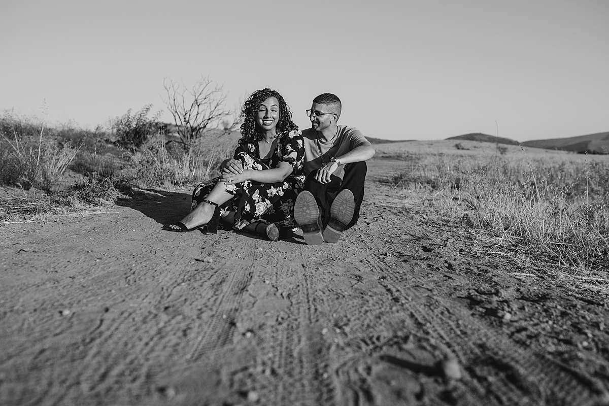 Orange County family photographer. candid black and white portrait of couple sitting on a dirt road while laughing at Irvine Regional Park Orange County
