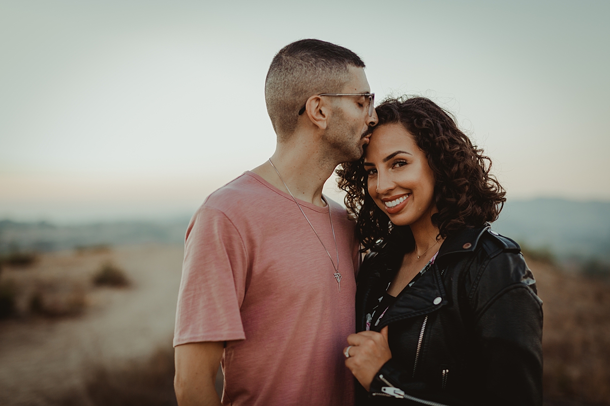 Orange County family photographer. photo of husband kissing wife while she smiles at camera wearing leather jacket