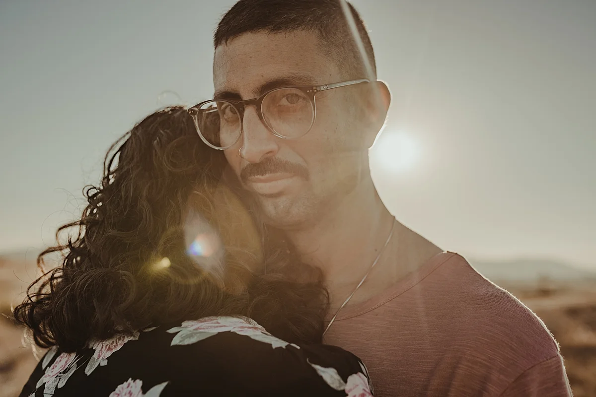 orange county family photographer. portrait of couple embracing while husband looks at the camera with sun flare in the background at Irvine Regional Park Orange County