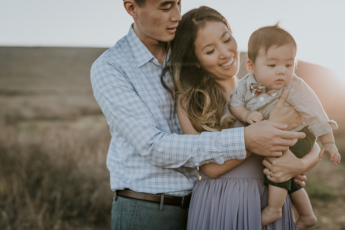 Orange County family photographer. Backlit photo of mom and dad holding their baby boy in a field of yellow grass during lifestyle family photo shoot at Quail Hill Trailhead in California