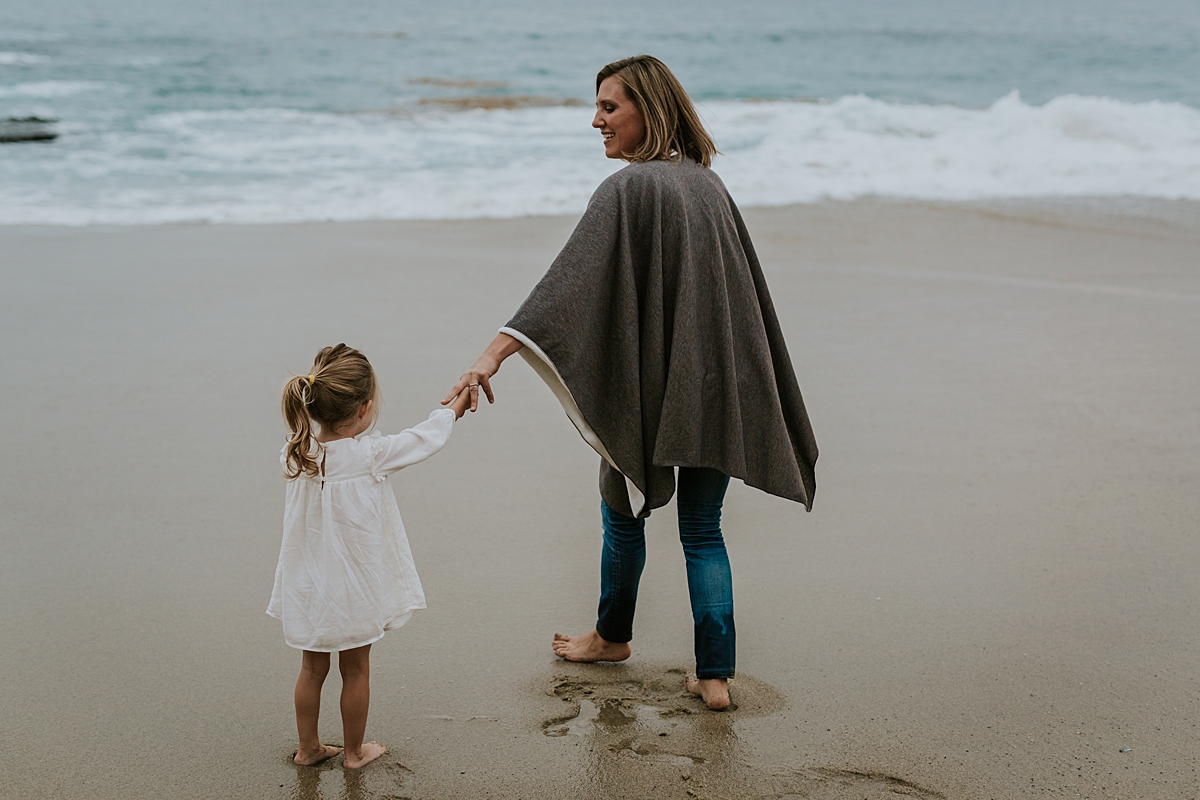 Orange County family photographer. Mom and her two year old daughter stand hand-in-hand in front of the waves at Table Rock Beach, Laguna Beach