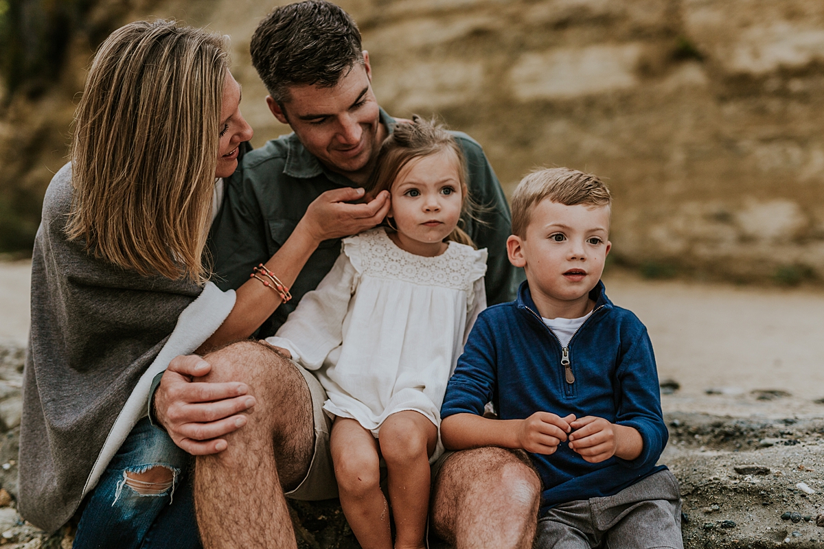 Orange County family photographer. Family of four sit in front of bluffs at Table Rock Beach Laguna Beach during photo session with Krystil McDowall Photography
