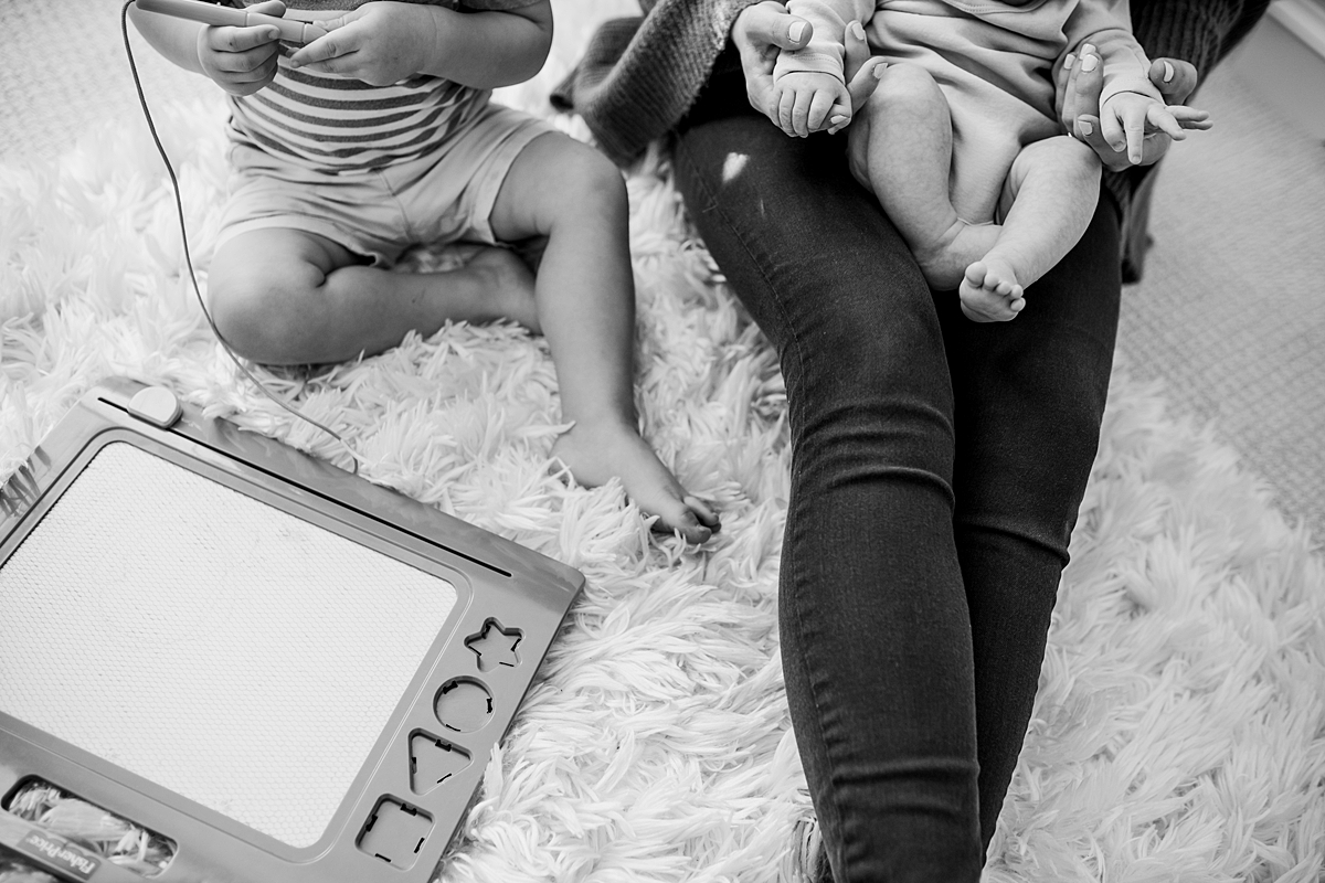 Orange County family photographer. Mom, toddler and newborn sit together playing in baby room during in-home newborn session with Krystil McDowall Photography
