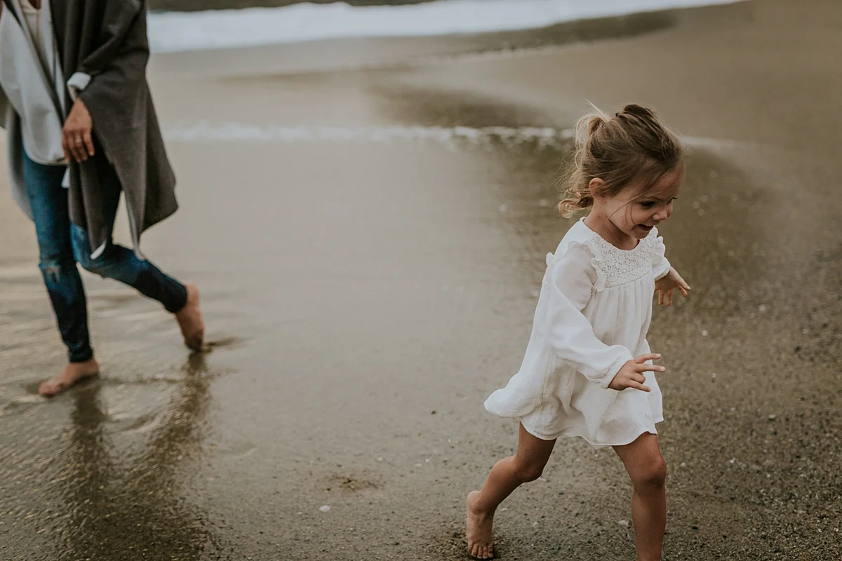 Orange County family photographer. Image of young girl in white dress running along beach away from her mom
