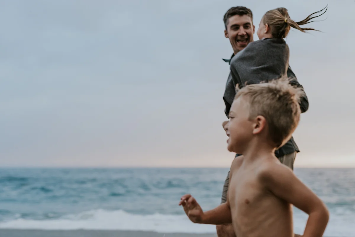 Orange County family photographer. Candid photo of dad running along the beach with his 2 year old daughter and son