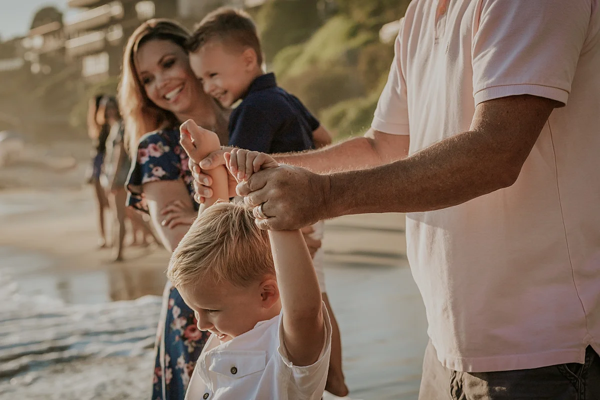 Orange County family photographer. Sunlit photo of family of four playing in shallow ocean waves at Corona Del Mar Beach during family photo session