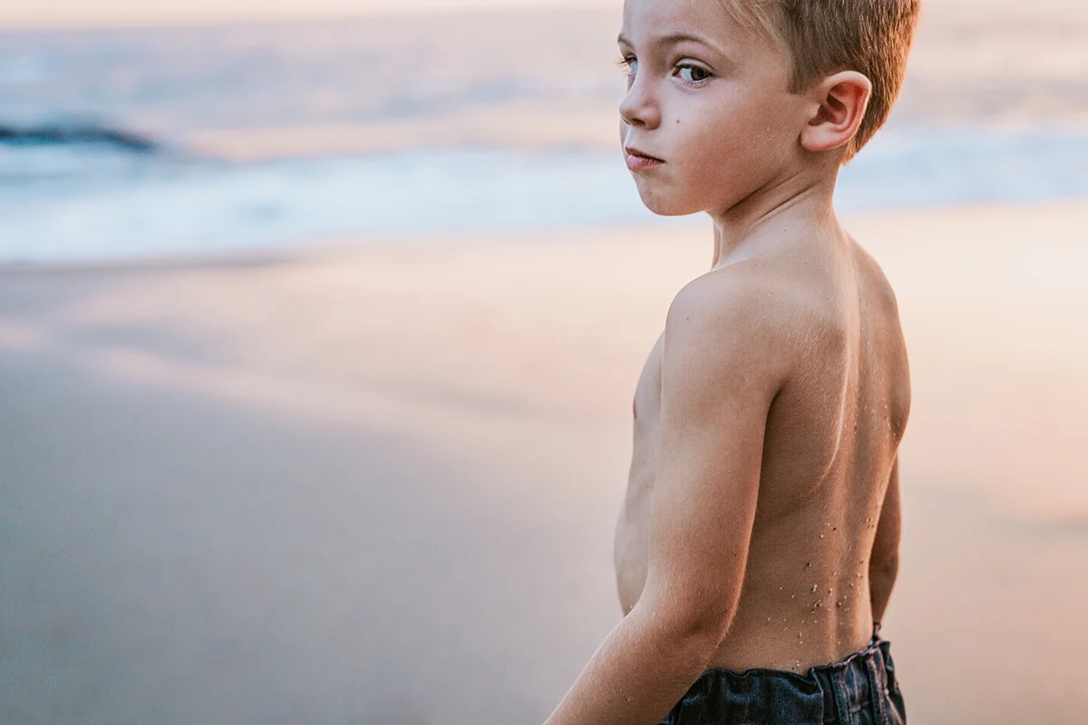 Orange County family photographer. Photo of young boy standing in the sand with pink sunset lighting up the sky during outdoor family photo session at Table Rock Beach Laguna Beach