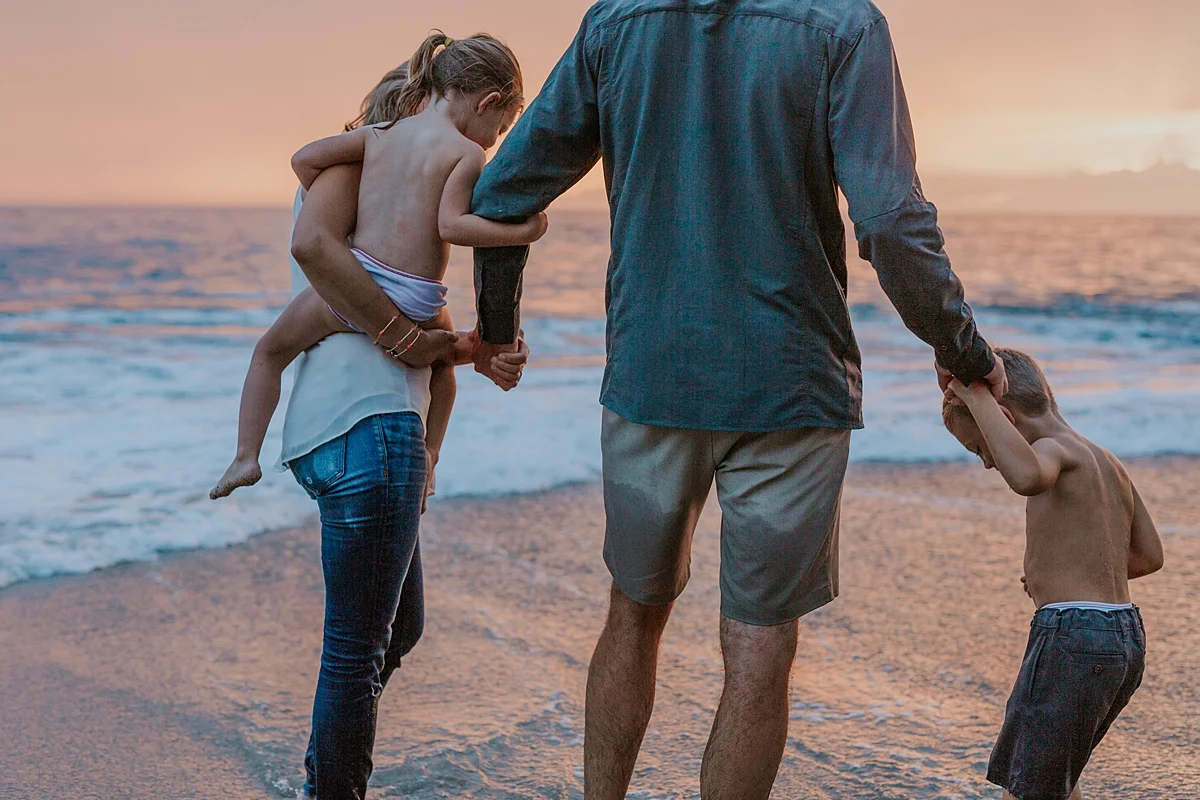 Orange County family photographer. Photo of family of four standing in the shallow ocean waves with pink sunset in the background during outdoor family photo session at Table Rock Beach Laguna Beach