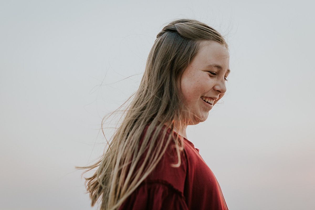 Orange County family photographer. Portrait of girl in maroon dress with long blonde hair streaming in the sunset during candid family photo session at Top of the World Laguna Beach