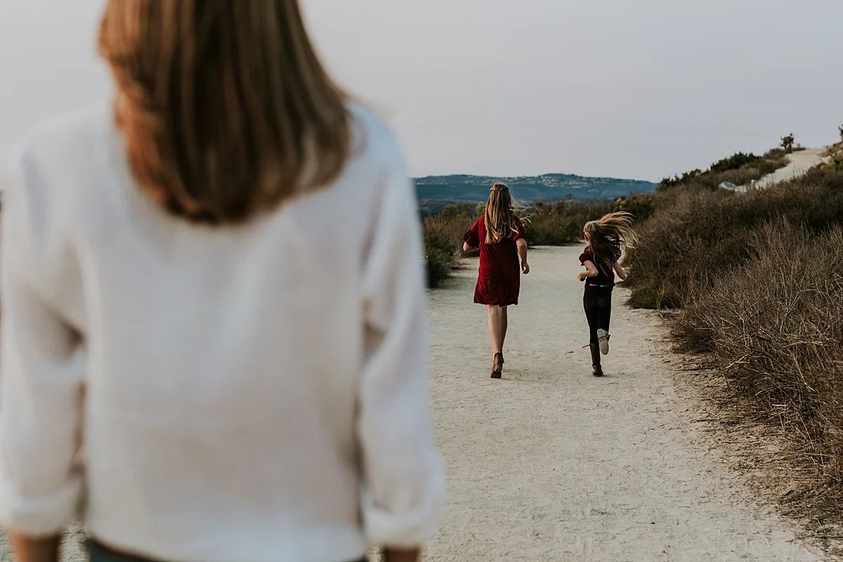 Orange County family photographer. Two sisters running along dirt road as mom looks on during family photo session at top of the world