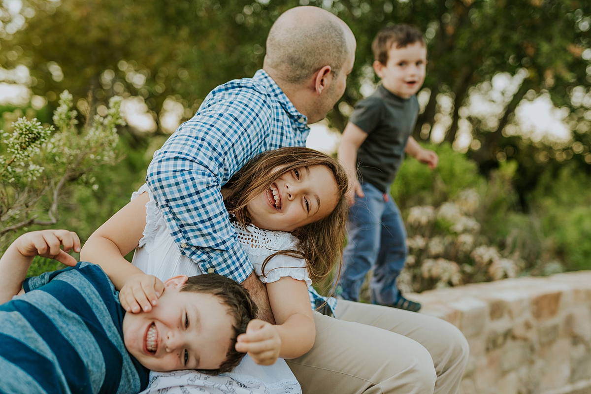 Orange County family photographer. Candid photo of dad and his 3 kids sitting on stone wall and playing together during family photo shoot at Quail Hill with Krystil McDowall Photography