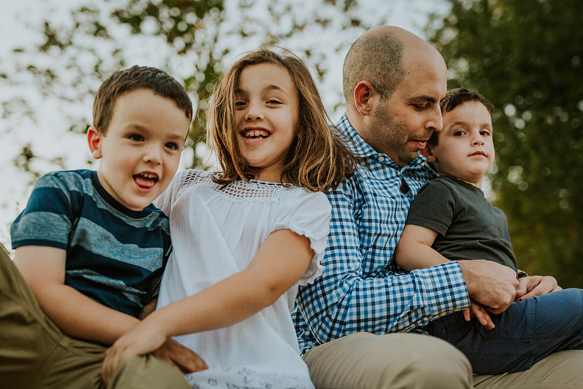 Orange County family photographer. Candid photo of dad and his 3 kids sitting on stone wall and playing together during family photo shoot at Quail Hill with Krystil McDowall Photography