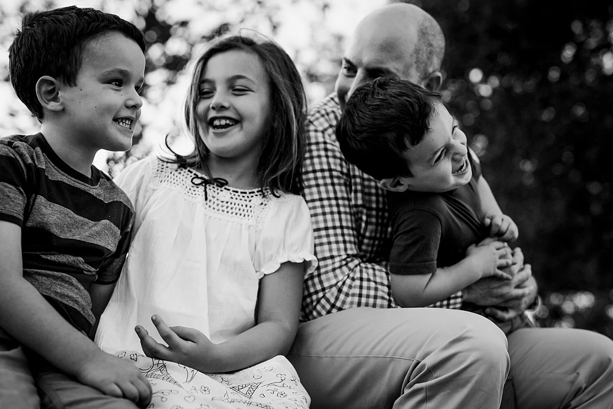 Orange County family photographer. Candid photo of dad and his 3 kids sitting on stone wall and playing together during family photo shoot at Quail Hill with Krystil McDowall Photography