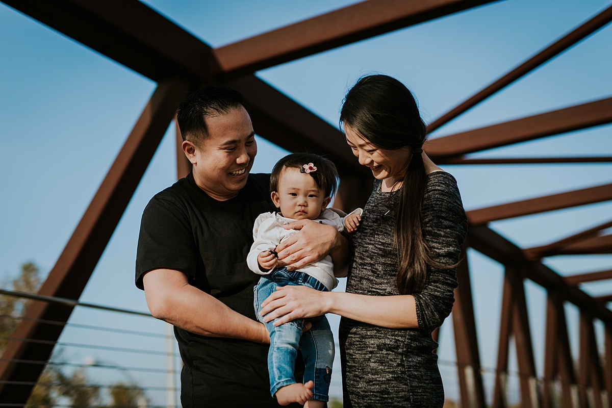 Orange County family photographer. Photo of mom, dad and sweet one year old daughter standing on bridge during outdoor family photo session at Jeffrey Open Space Trail in Irvine