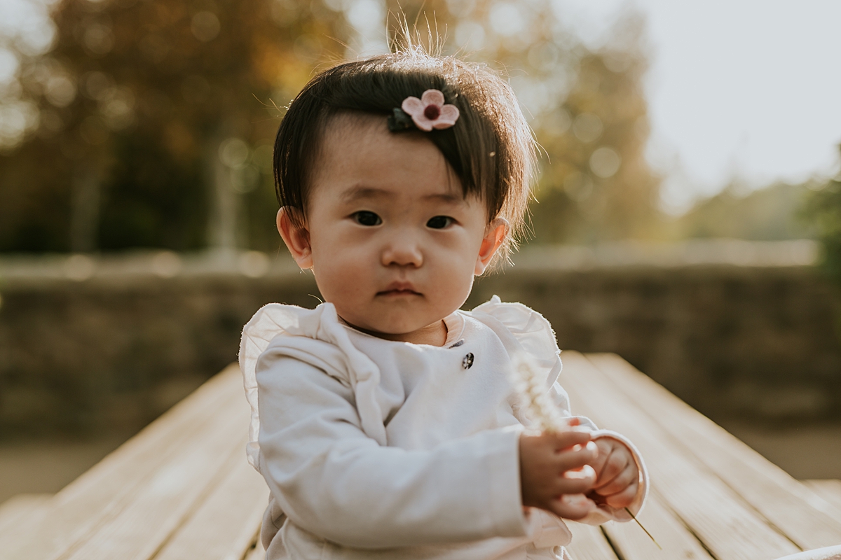 Orange County family photographer. Portrait of one year old girl with pink bow sitting on park bench during outdoor family photo session at Jeffrey Open Space Trail Irvine