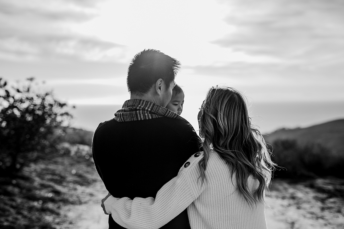 Orange County family photographer. Black and white backlit photo of mom, dad and daughter walking into the sunset during during outdoor during family photo session at Top of the World Laguna Beach