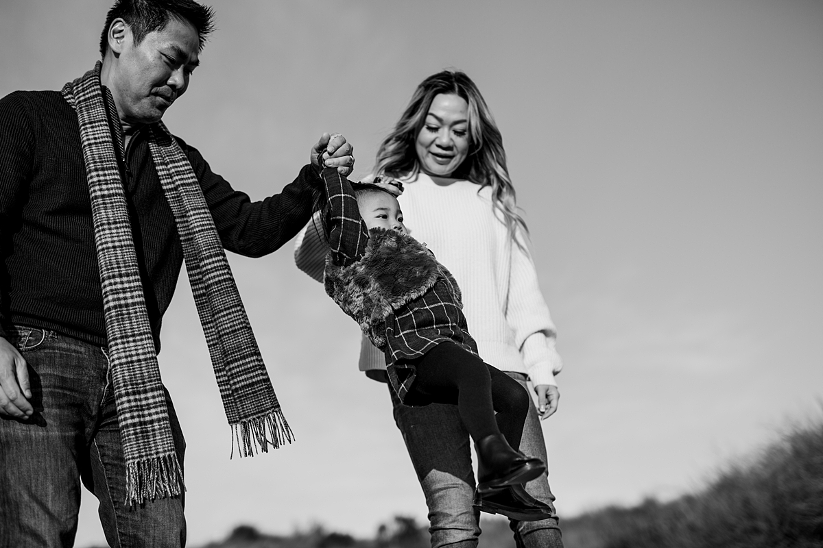 Orange County family photographer. Photo of toddler being swung in the air by her mom and dad during outdoor during family photo shoot at Top of the World Laguna Beach CA