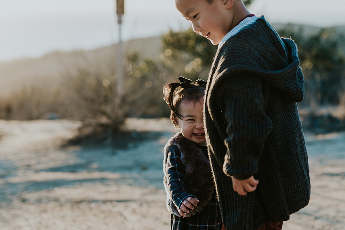 Orange County family photographer. Photo of brother and sister hugging while playing on a dirt road in the sun during outdoor during family photo shoot at Top of the World Laguna Beach CA