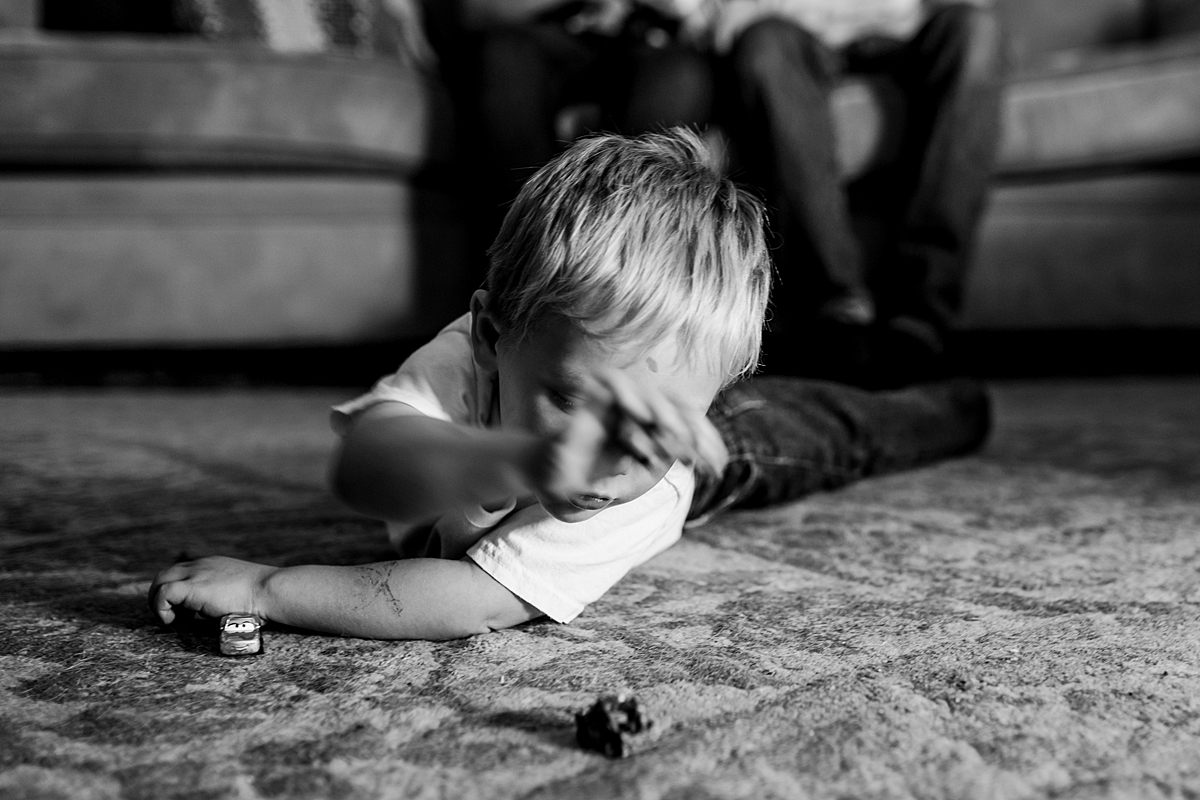 Orange County family photographer. Black and white candid photo of toddler playing with cars on the carpet while mom, dad and newborn son hang out in the background during in home newborn session