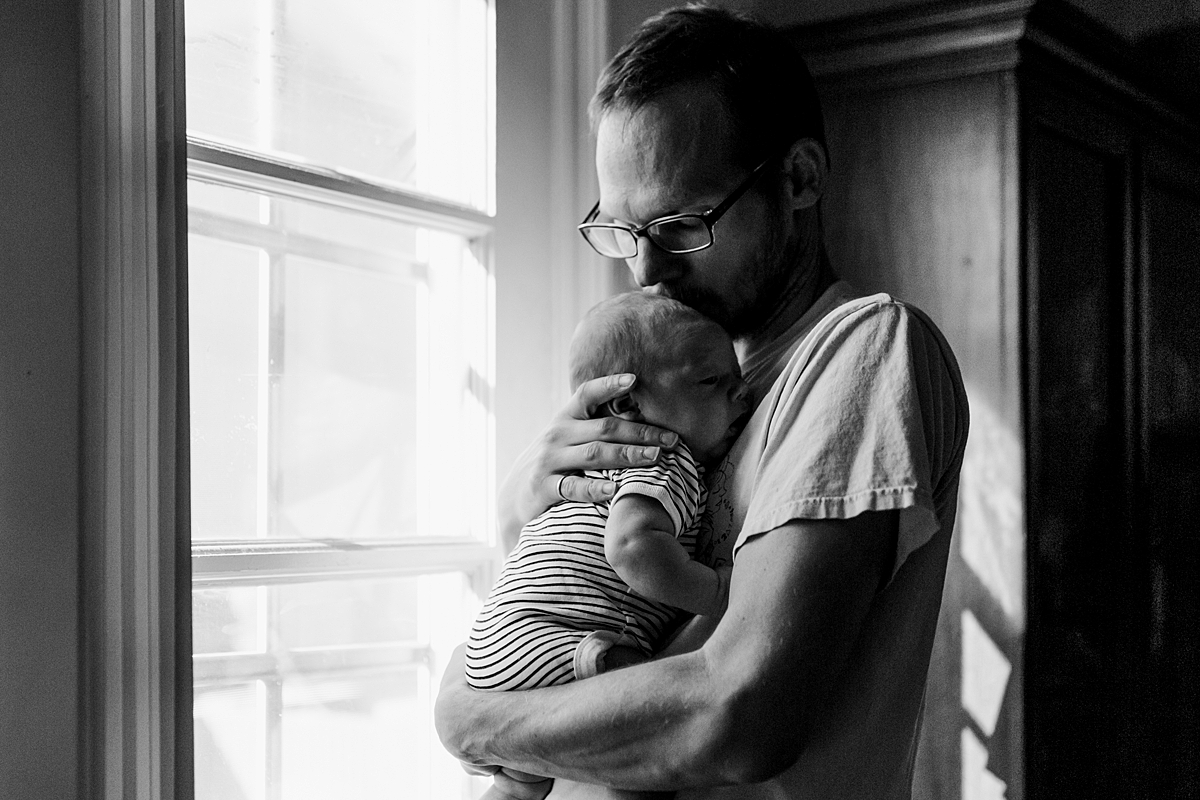 Orange County family photographer. Black and white photo of dad and his newborn son as they stand near light filled window during in home newborn session with Krystil McDowall Photography