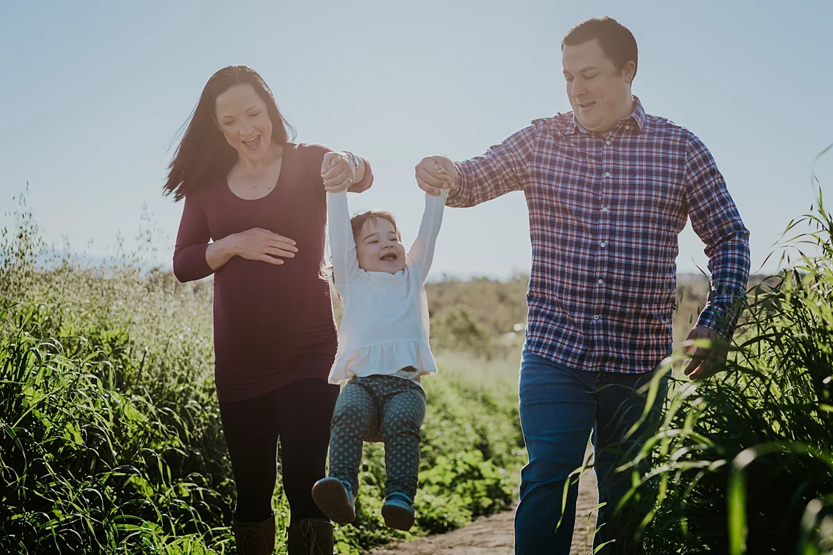Orange County family photographer. Photo of expecting mom and dad swinging their toddler daughter in the long green grass during maternity shoot at Quail Hill Trailhead Irvine