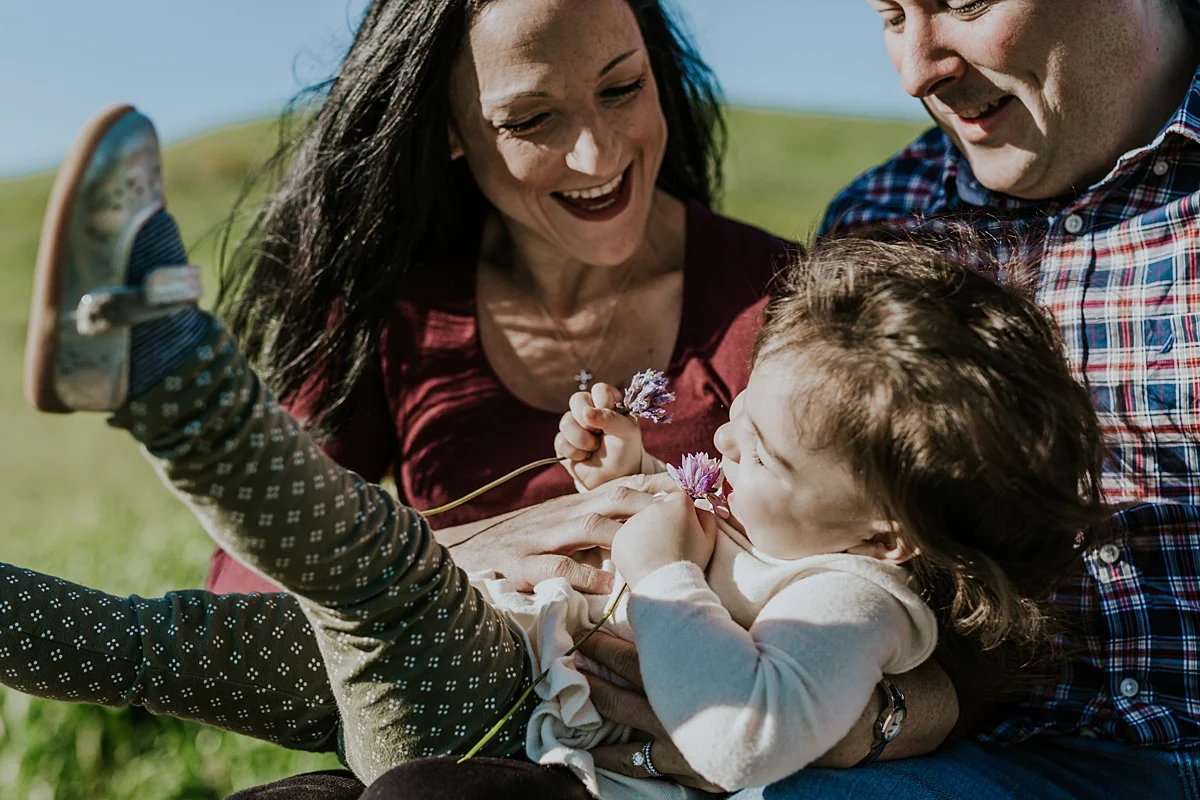 Orange County family photographer. Candid photo of mom and dad tickling baby girl as she lays on their lap during maternity photo shoot at Quail Hill Trailhead Irvine