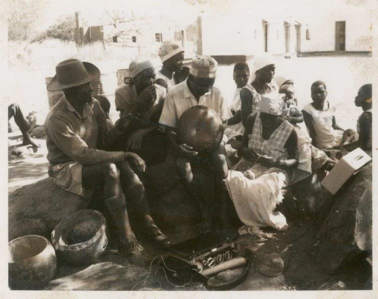 ILAM Photograph collection #ILM00426 381_Johani holds karimba while listening to playback_1969. Seated next to him on the right is Sinati Kadende (known these days as Sinati Nyamande) and seated behind her, also in the white headwrap, is Mavis Bandeira. 