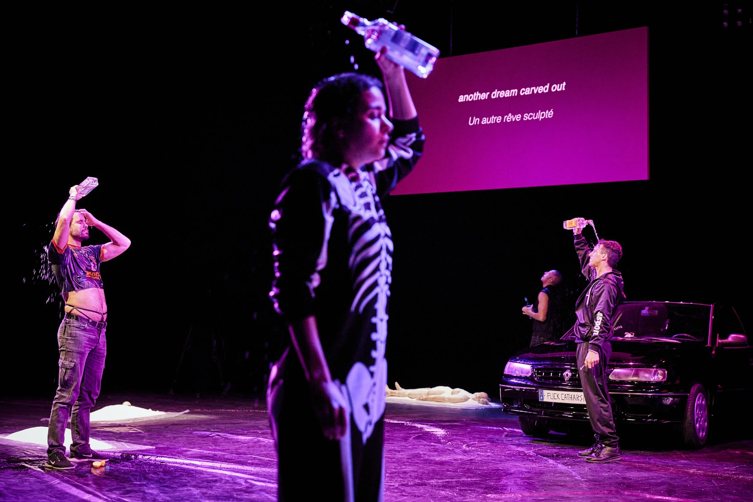  Three people on a dark stage around a car pour alcohol over their heads while a body lies on the floor. The words 'another dreamed carved out' and below that 'Un autre rêve sculpté' are projected onto a screen.  