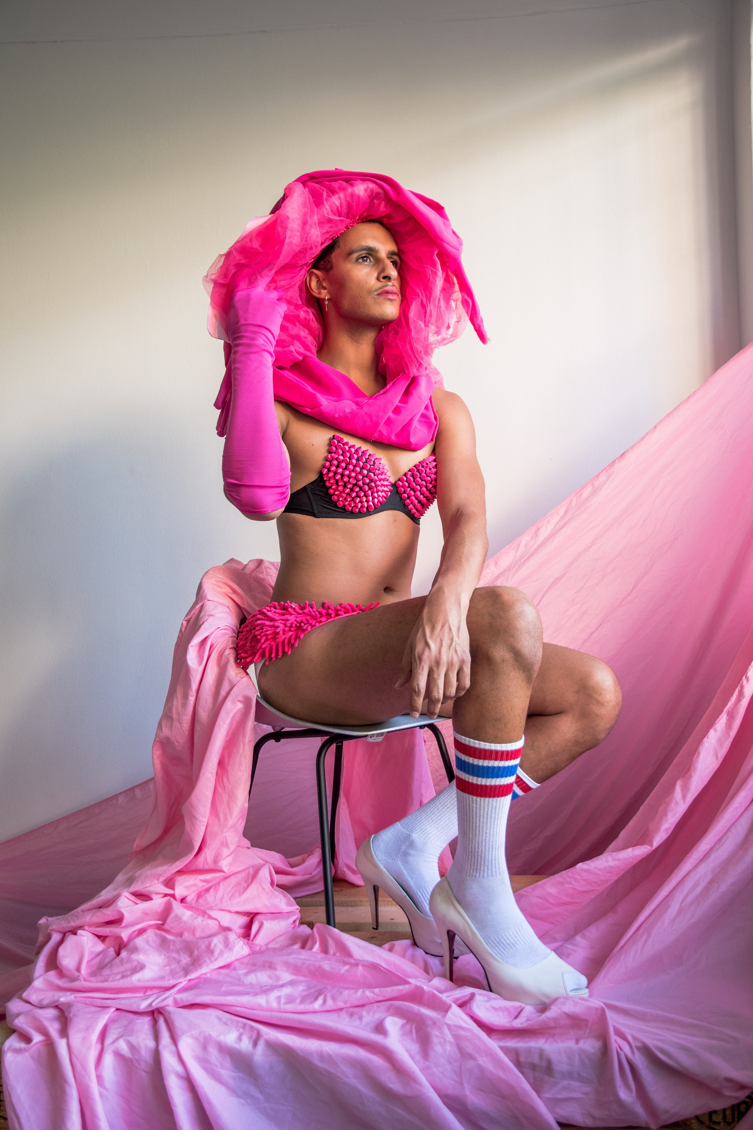  This is an image of Kieron Jina. Kieron is posed sitting on a chair with a white wall behind him that has a pink sheet draped behind them. He is wearing a large matching bright pink hat, one long glove, a black and pink bra, pink underwear, white hi