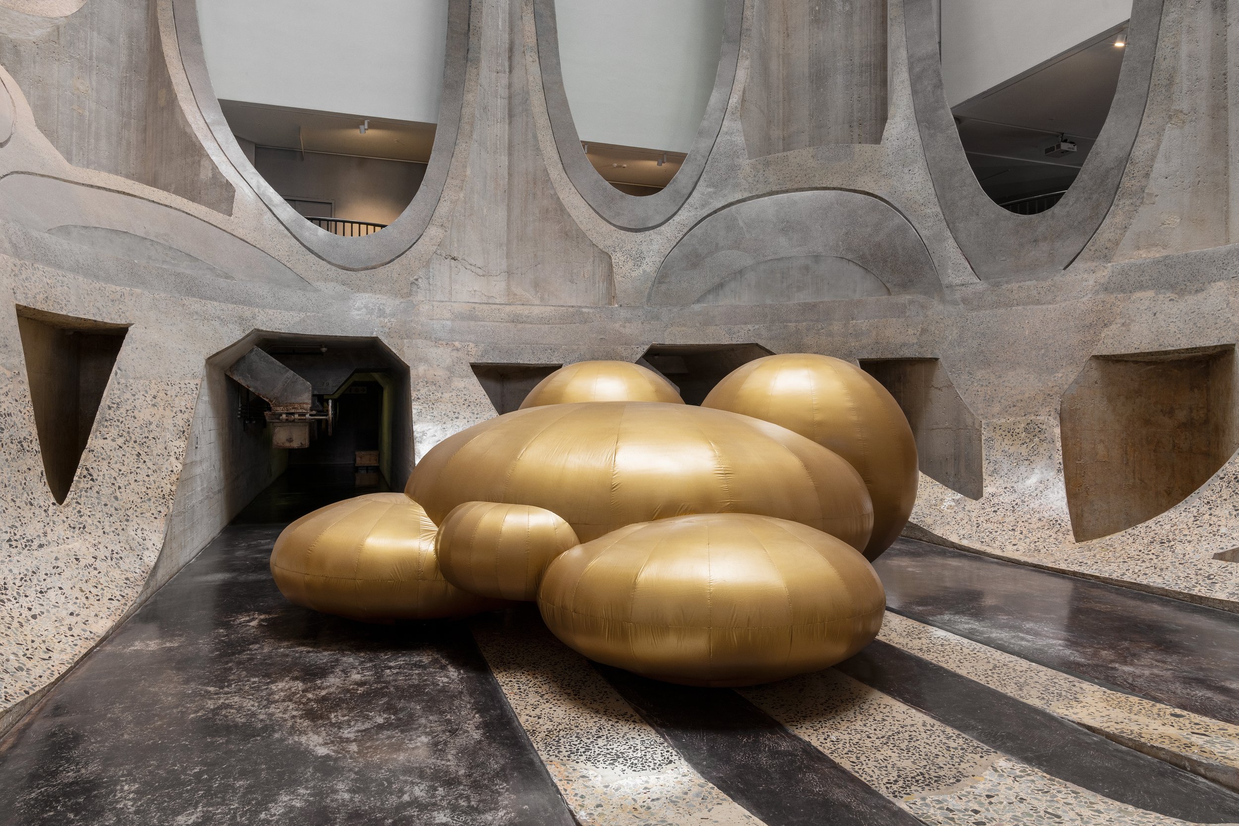  This image shows GoldenDean's Soft Vxnxs a large interactive inflatable sculpture. The sculpture is gold, and made of round shapes that altogether resemble a human figure. It is situated in a large concrete space. 